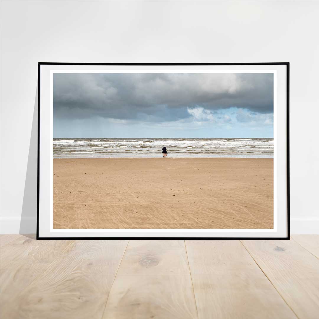 This photograph of a lone woman standing on an empty beach with her back to the camera, facing a receding tide under large, dark clouds, evokes a profound sense of solitude and introspection.