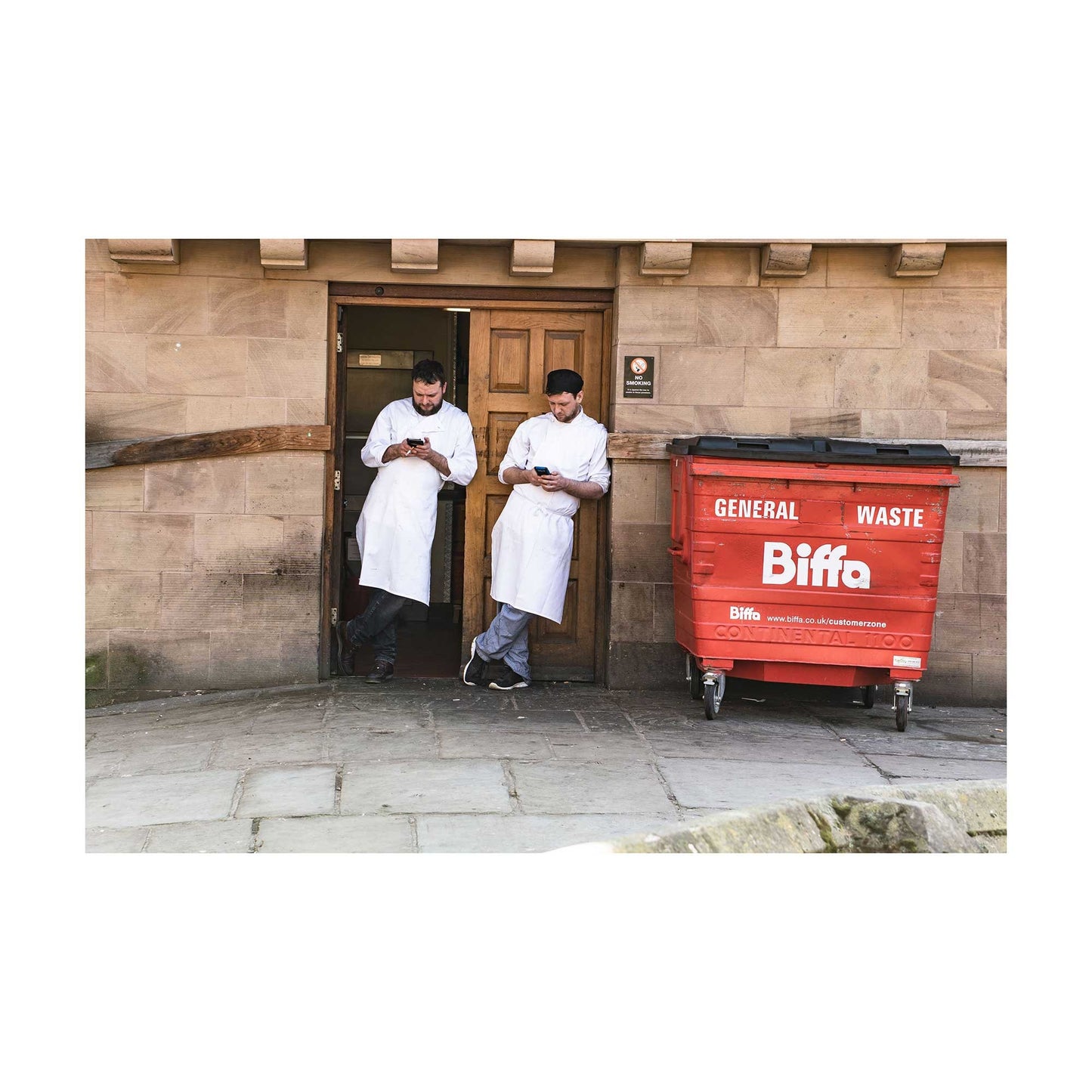 A photographic print was taken in Manchester, England showing street photography of two men on a break who stood by a wall.